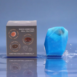 Jabón elemental cristal agua en forma cúbica azul translúcido junto a caja gris con gemas incrustadas y texto promocional, ver r
