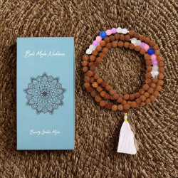 Collar Bali Mala de cuentas marrones con cuentas rojas, abalorios morados y rosados, y colgante de borla blanca junto a una caja